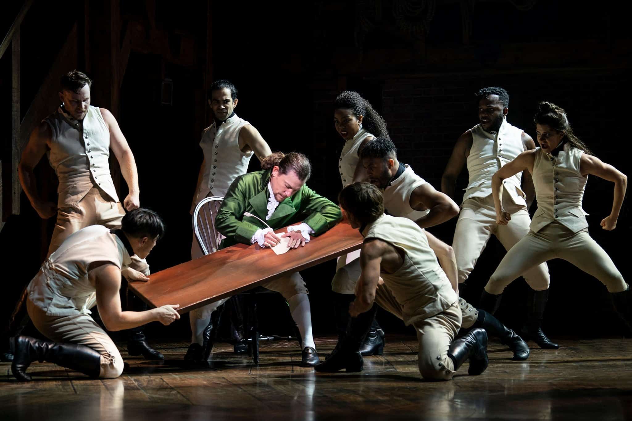 A dramatic scene from the Hamilton musical with a performer in a green coat writing on a tilted desk, surrounded by energetic dancers in beige costumes.